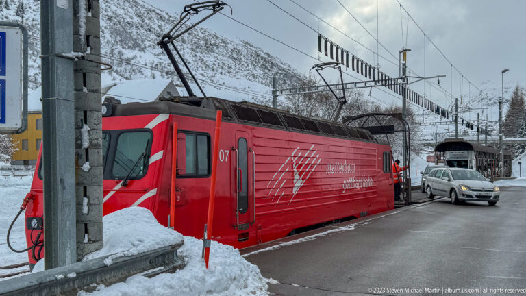 Close side view of a red Matterhorn Gotthard Bahn locomotive beneath catenary wires, with a silver car alongside.
