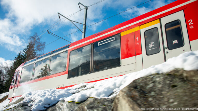 Red-and-white electric passenger train passing above a snowy embankment under overhead catenary wires.