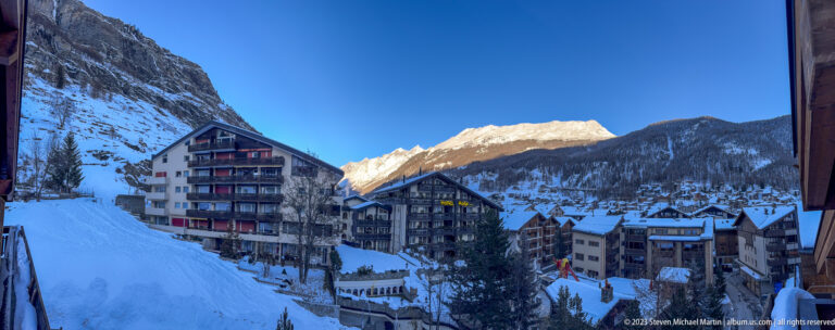 Snow-covered Zermatt street and multi-story hotels in blue shade beneath sunlit mountain peaks.