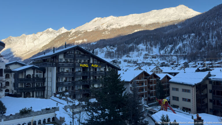 Hotel Alex and neighboring snow-covered buildings in Zermatt beneath a brightly sunlit mountain ridge.