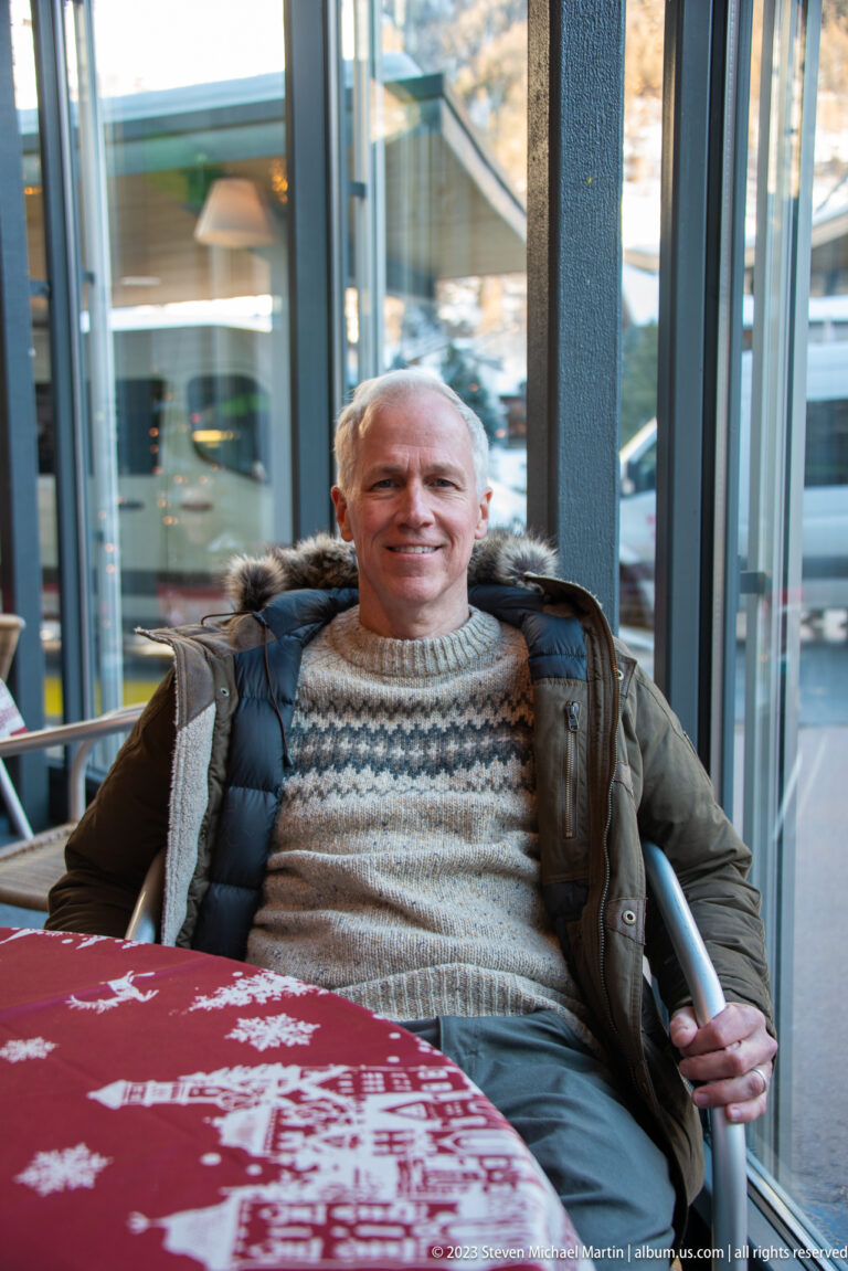 Man in patterned sweater and winter coat seated at a red-covered table beside large windows.