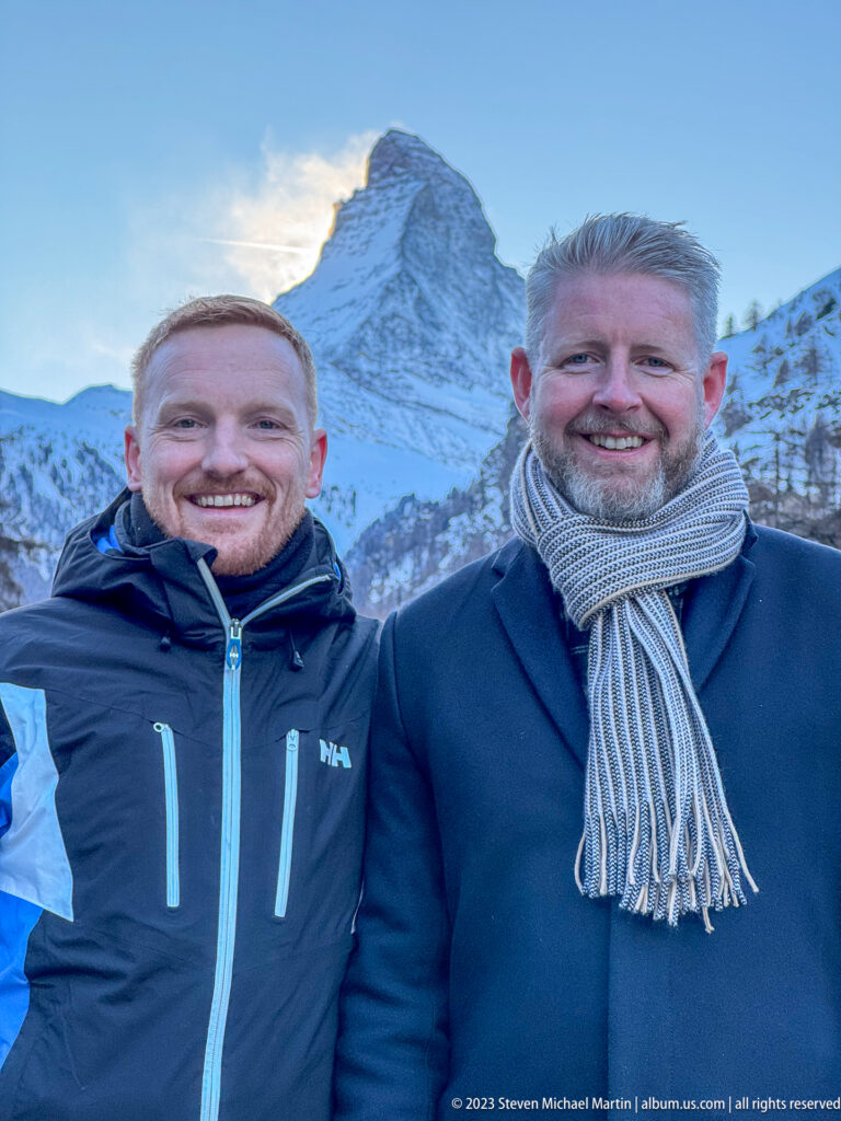 Two smiling men in winter coats posing outdoors with the snow-covered Matterhorn directly behind them.