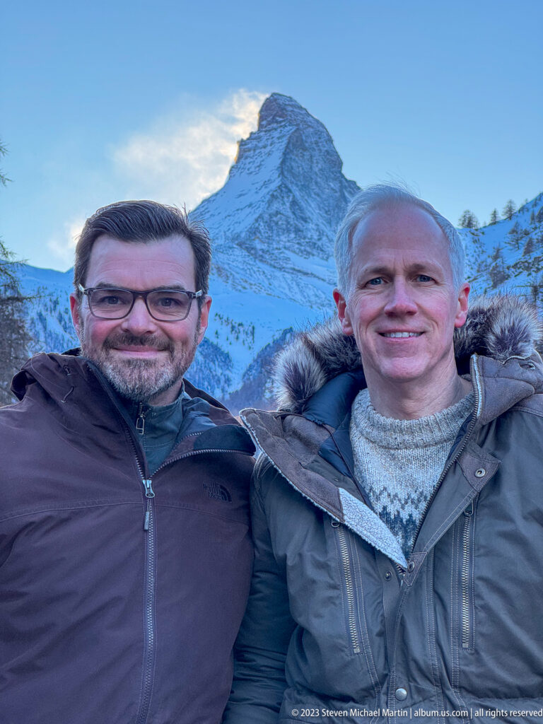 Two men in winter coats posing close to the camera with the Matterhorn rising behind them.