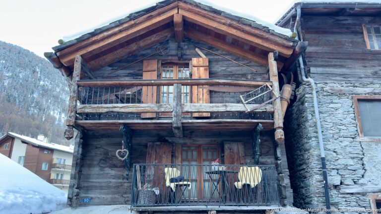 Weathered two-story timber chalet with stacked balconies and rough wooden railings beside a stone-walled building.