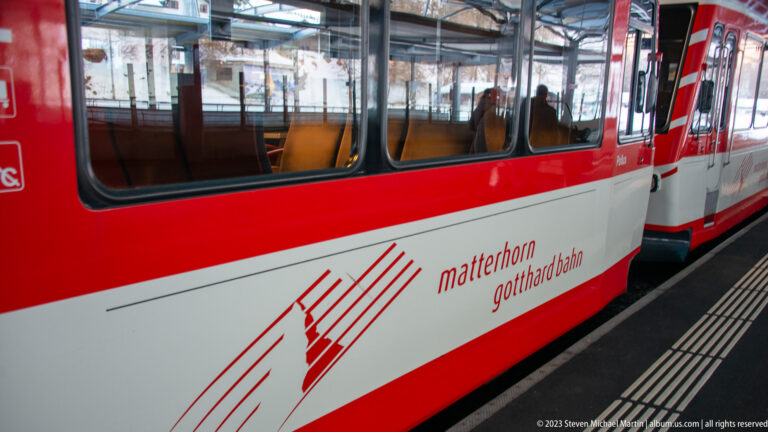 Red-and-white Matterhorn Gotthard Bahn train beside a station platform, with a second railcar visible beyond.