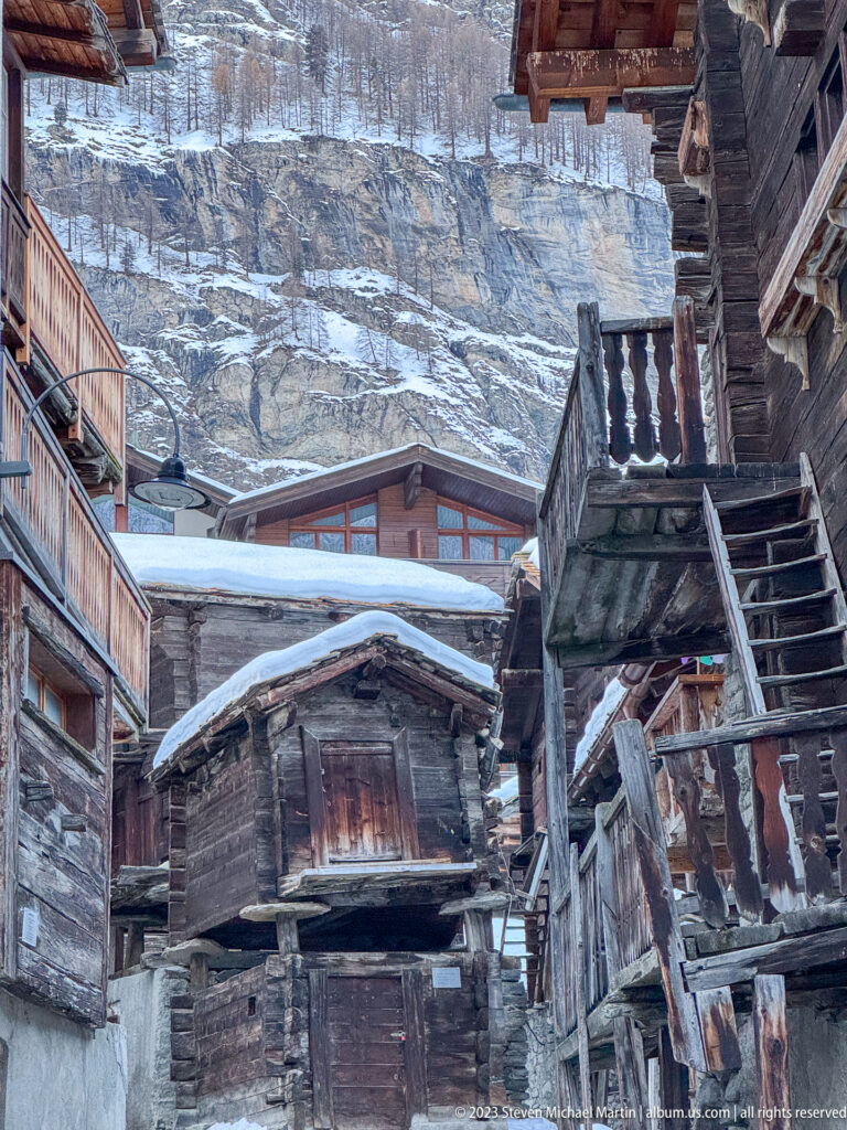 Cluster of weathered timber buildings with exterior stairs, balconies and snowy roofs against a rocky slope.
