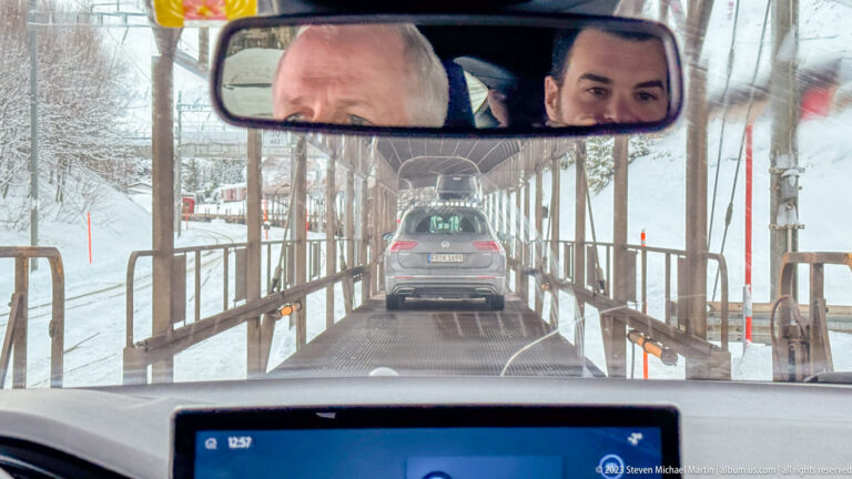 View through a windshield of a car ahead in a narrow covered passage, with snowy slopes outside and two faces reflected above.