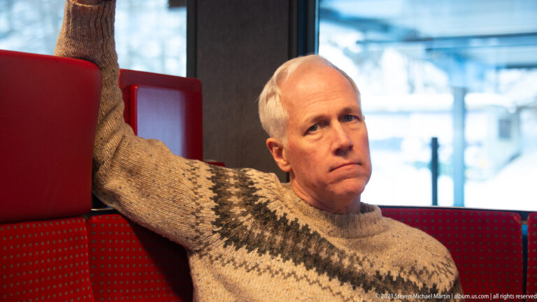 Man in a patterned wool sweater seated against a red train seat with one arm raised along the backrest.