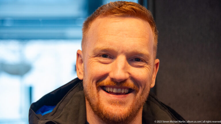 Close-up portrait of a smiling red-haired man indoors against a softly blurred background.