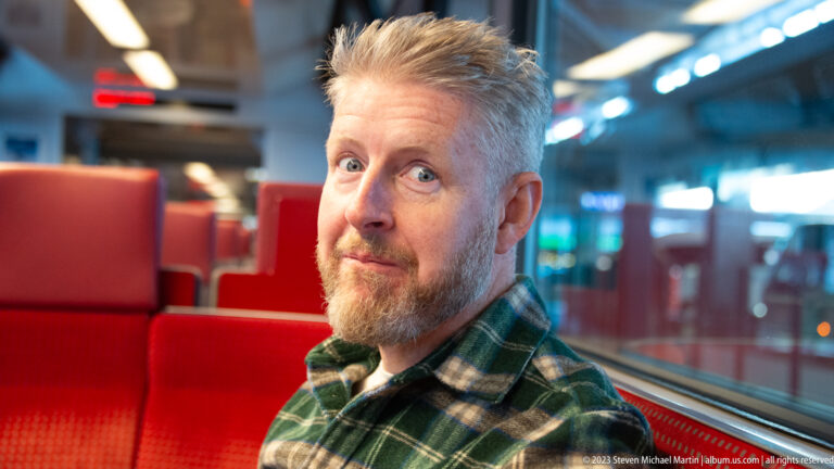 Gray-haired bearded man in a green plaid shirt seated beside a train window and red seats.