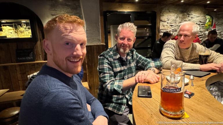 Three men seated at a wooden restaurant table with a large glass of Cardinal beer in the foreground.