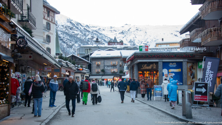 Pedestrians walking along a shop-lined street in Zermatt with snowy mountain slopes rising beyond the buildings.