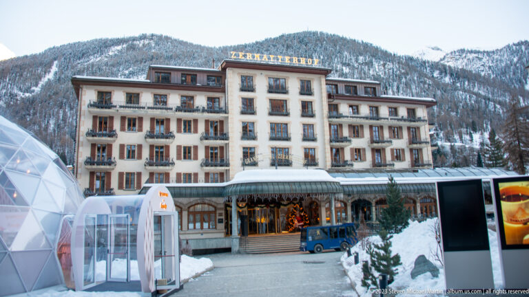 Zermatterhof hotel with its name on the roofline, symmetrical facade, and snowy mountain slopes behind.