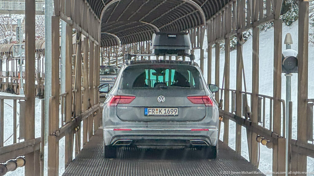 Gray Volkswagen with roof box driving through a narrow open-sided covered passage surrounded by snow.