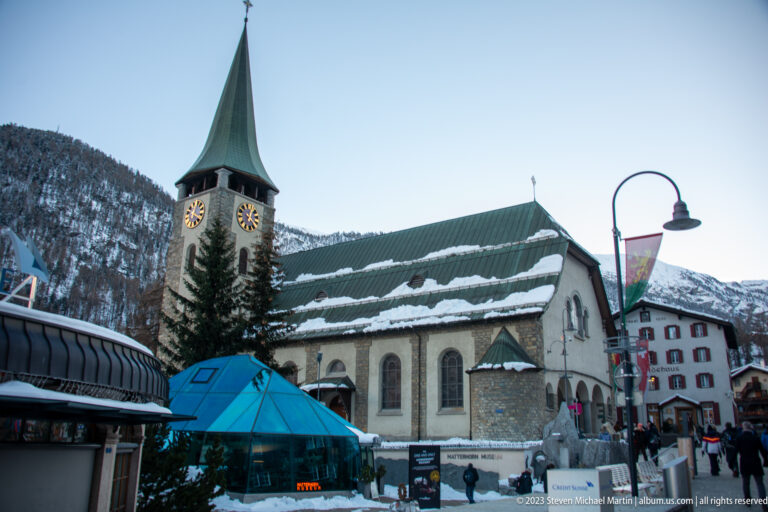 Stone Parish Church of St. Mauritius with clock tower, green spire, and snow-covered roof in Zermatt.