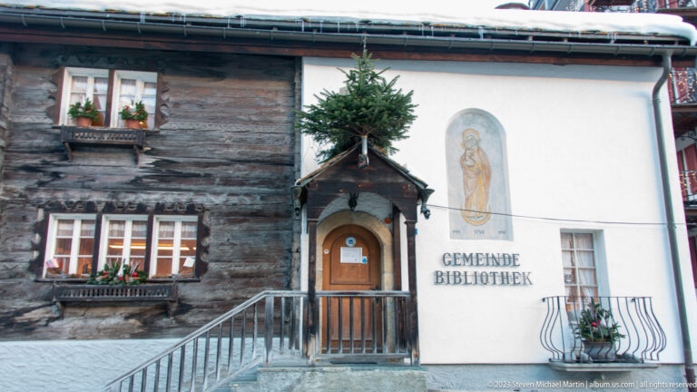 Historic timber-and-plaster building marked GEMEINDE BIBLIOTHEK with a small gabled doorway and religious wall painting.