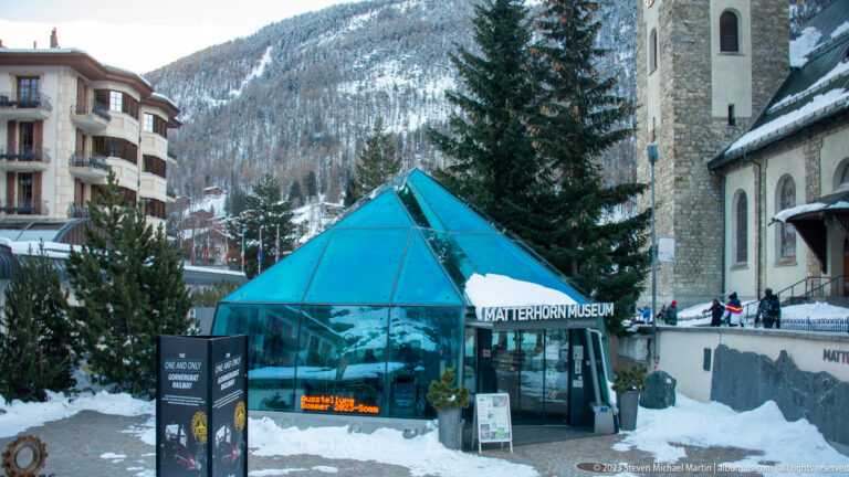 Blue glass Matterhorn Museum entrance pavilion in snow beside the stone parish church.