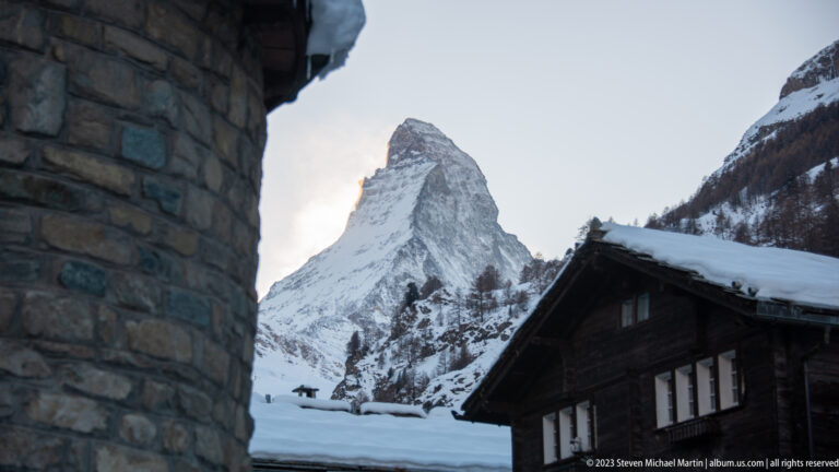 Matterhorn framed between a stone building and dark timber chalet, with snowy roofs below.