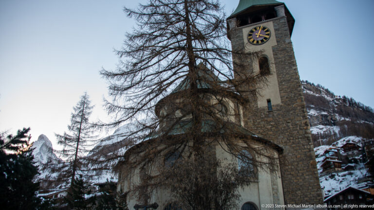 Stone clock tower of St. Mauritius Church behind bare larch branches, with a glimpse of the Matterhorn at left.