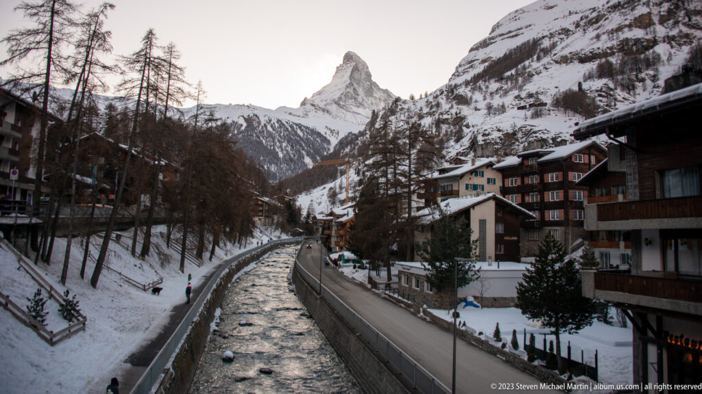 Matter Vispa flowing through Zermatt between snowy banks and buildings, with the Matterhorn in the distance.