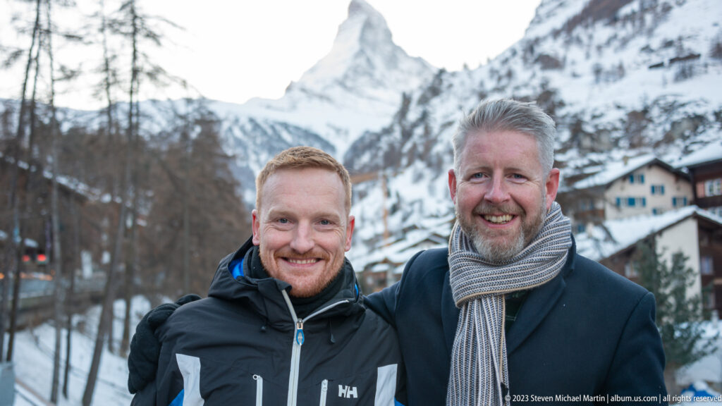 Two men in winter clothing posing beside the Matter Vispa with the Matterhorn centered behind them.