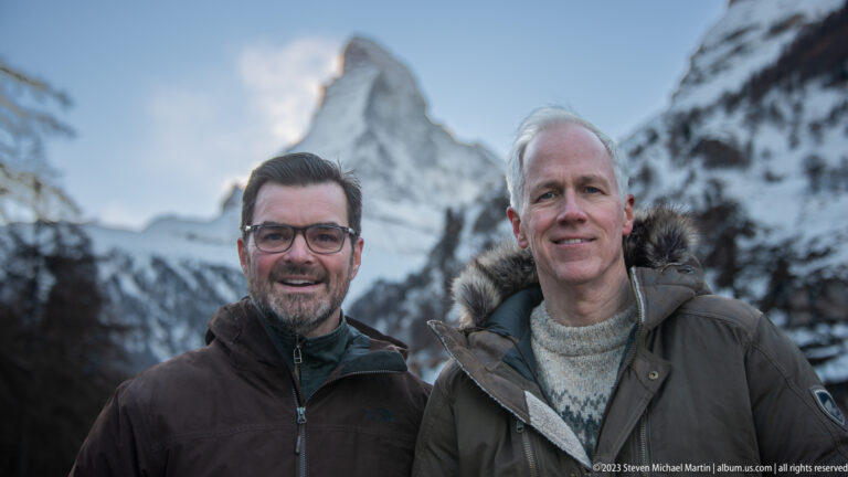 Two men in winter coats posing close to the camera with the Matterhorn softly focused behind them.