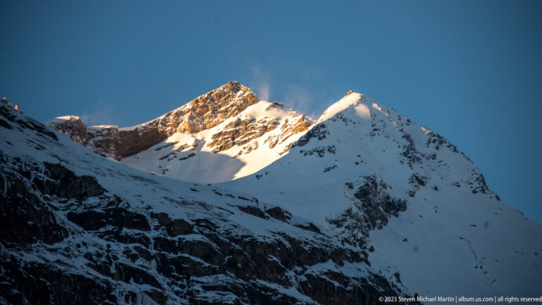 Sunlit snow-covered mountain summit above dark shadowed slopes, with windblown snow streaming from the ridge.