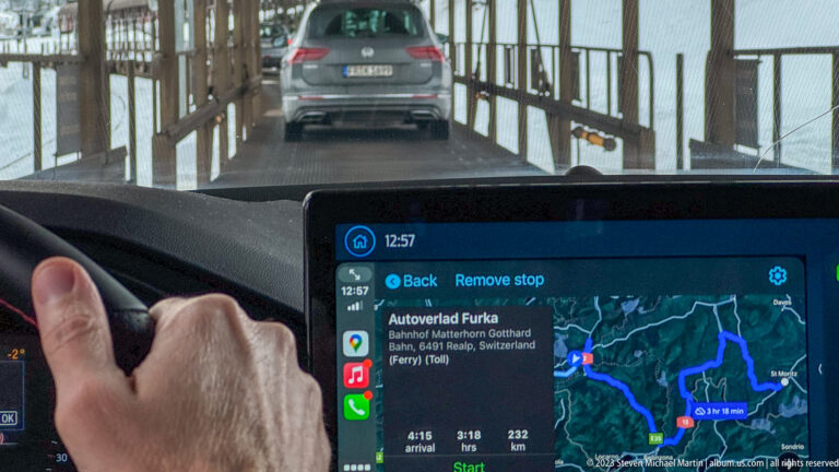 Driver's hand and navigation screen showing Autoverlad Furka directions while following a car through a covered passage.