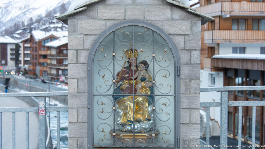 Gilded Madonna and Child statue behind an ornate iron grille inside a small arched stone shrine.