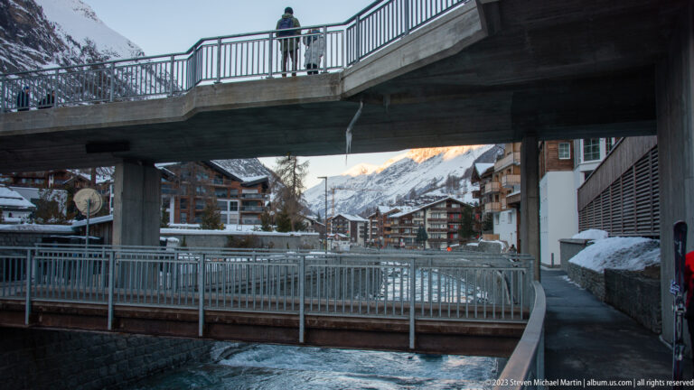 Pedestrian bridge crossing the Matter Vispa beneath a concrete road bridge in snowy central Zermatt.