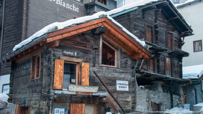Small dark timber house with wooden shutters and a steep snowy roof beside a taller historic building.
