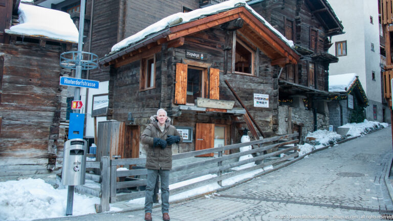 Man in winter coat standing beside historic timber houses on Hinterdorfstrasse in snowy Zermatt.