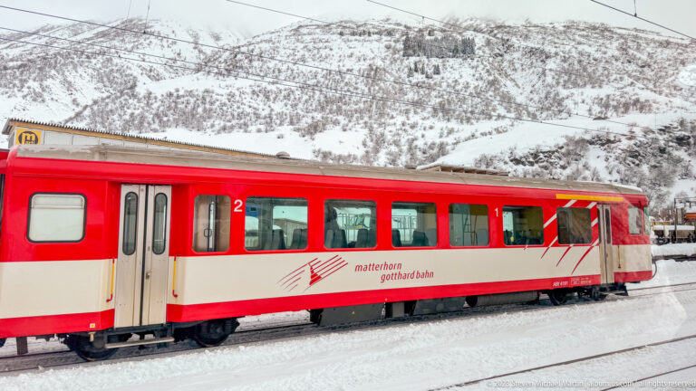 Red-and-cream Matterhorn Gotthard Bahn passenger railcar beside tracks below a snow-covered mountainside.
