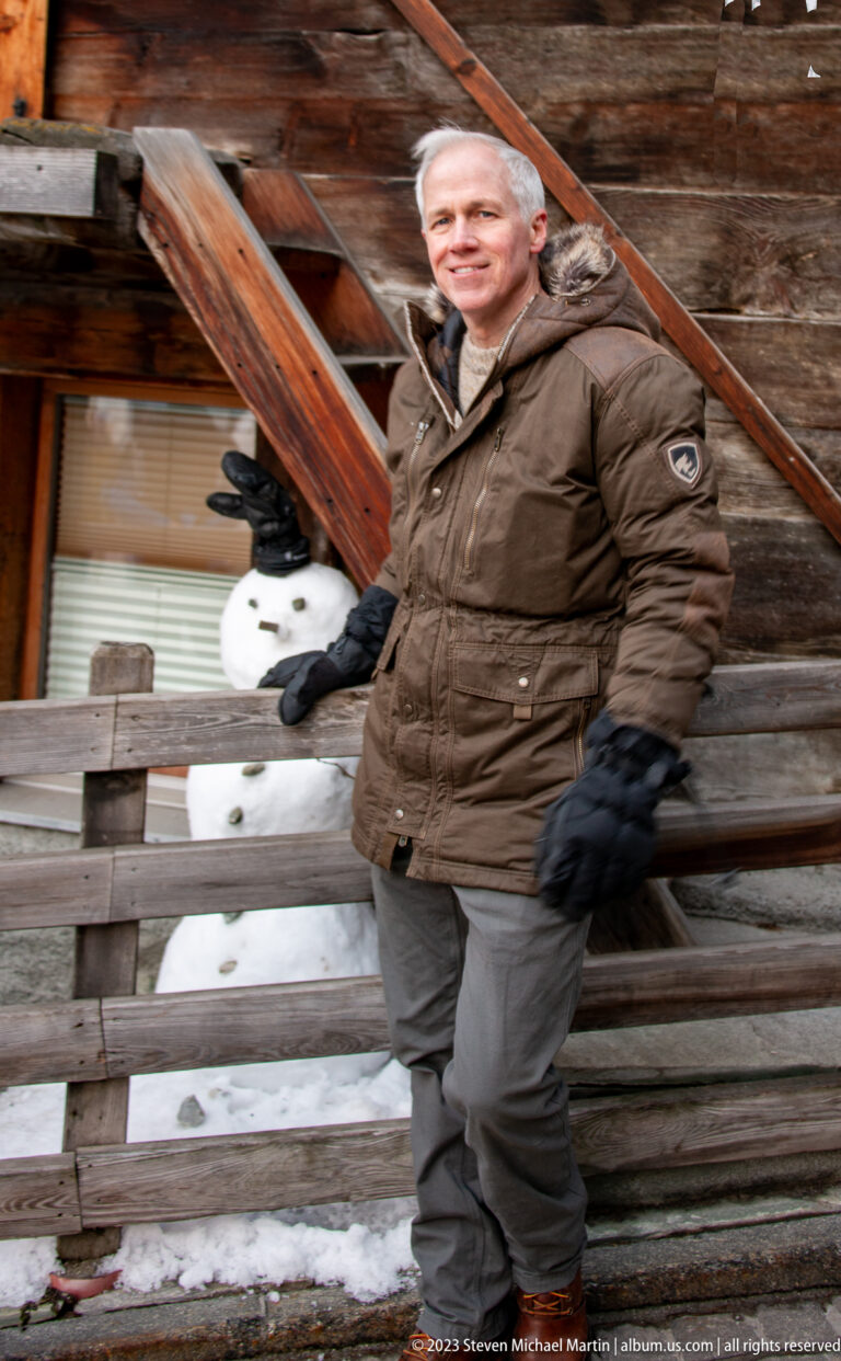 Man in a brown winter coat standing on wooden stairs beside a weathered timber building with snow.