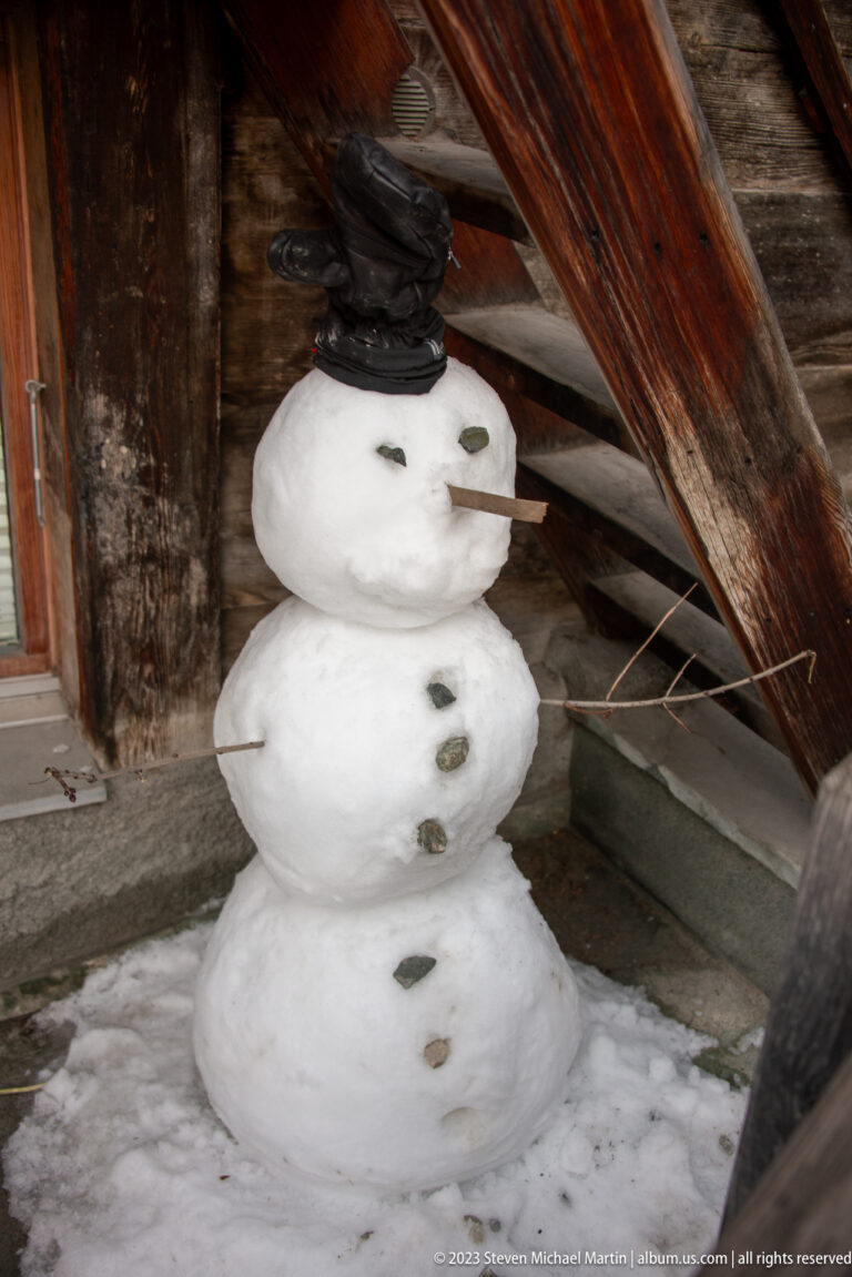 Small three-tier snowman with pebble buttons and twig arms beneath a weathered timber building.