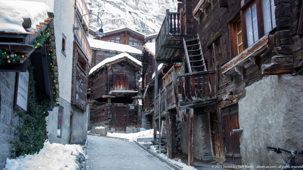 Narrow snowy lane between old timber houses with balconies and exterior stairs in Zermatt.