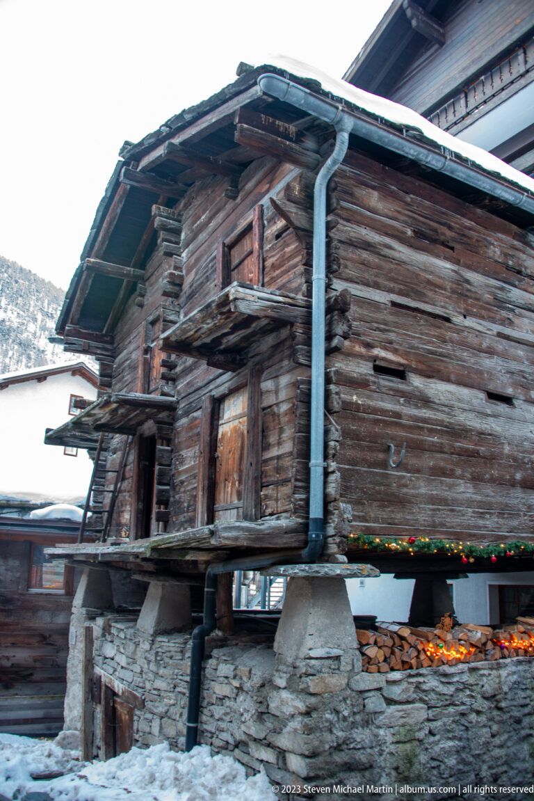 Weathered raised timber granary with broad roof, stone supports, and stacked firewood beneath.
