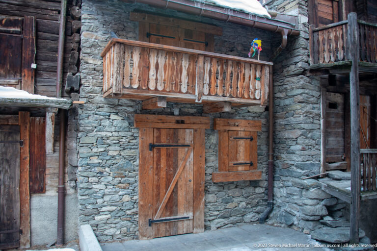 Stone-and-timber house with carved balcony, wooden door, and shutters in snowy Zermatt.