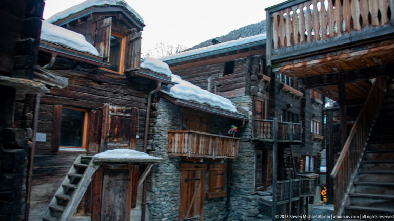 Clustered old timber and stone buildings with exterior stairs and balconies around a narrow snowy passage.