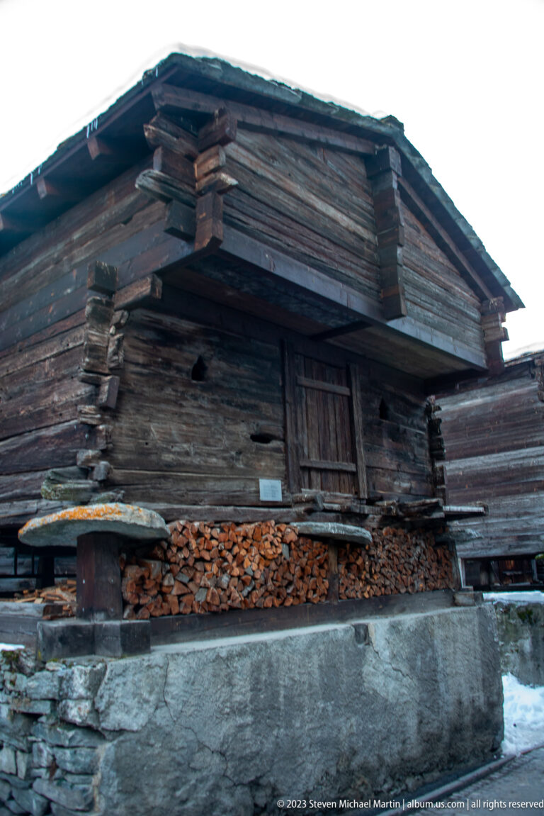 Weathered timber barn on a stone base with stacked firewood beneath its long overhanging eaves.