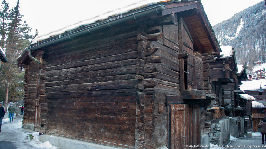 Large dark timber building with heavy log walls and steep roof beside a snowy pedestrian lane.