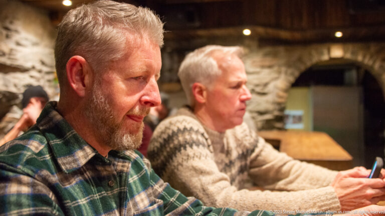 Two men seated at a wooden restaurant table against rough stone walls, wearing plaid and patterned sweaters.