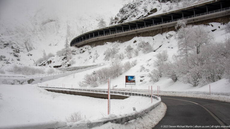 Cleared curving mountain road through deep snow below a long protective gallery on a steep Alpine slope.