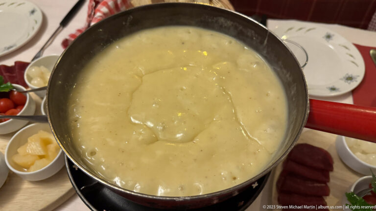 Red-handled fondue pot filled with melted cheese, surrounded by small bowls of accompaniments.