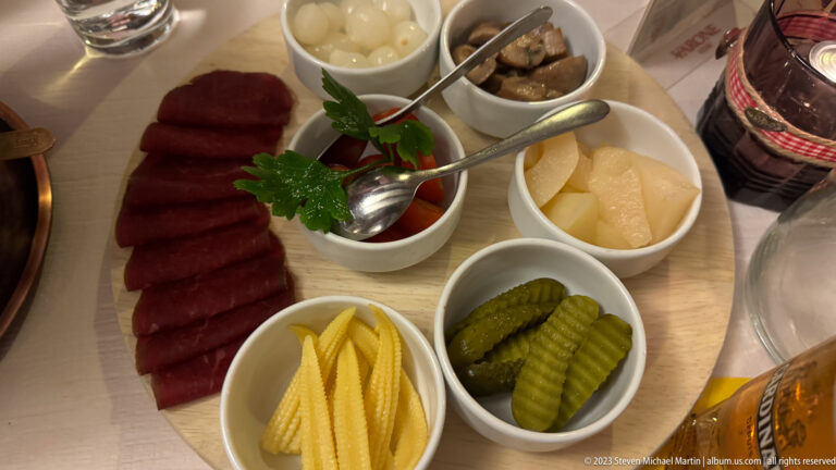Board with sliced cured meat and bowls of cornichons, baby corn, onions, mushrooms, tomatoes, and pale accompaniments.