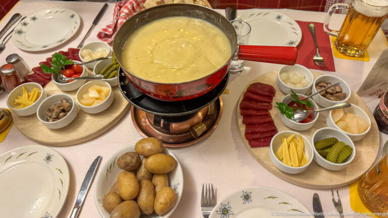 Cheese fondue pot on burner with boiled potatoes, charcuterie boards, pickles, vegetables, plates, and beer.