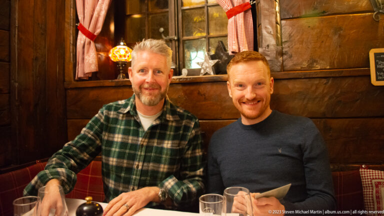 Two smiling men seated together at a restaurant table in a warm timber-lined interior.