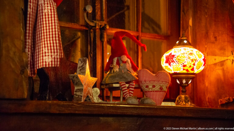 Red-hatted gnome beside rustic stars, a heart ornament, and a glowing multicolored lamp on a wooden shelf.