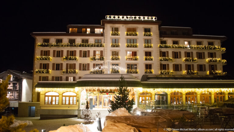 Grand Hotel Zermatterhof illuminated at night with balcony garlands and snow in the foreground.
