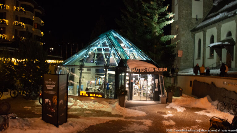 Illuminated glass Matterhorn Museum entrance pavilion at night beside snow and the church wall.